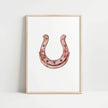 Elegant minimalist horseshoe in muted rose gold against white background, featuring detailed rivets and rustic finish, framed in natural wood for good luck charm in modern farmhouse decor.