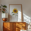 Watercolor sunflower in vibrant golden-yellow with delicate green leaves against a clean white background, displayed in a rustic wooden frame creating a minimalist, calming aesthetic.