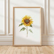 Watercolor sunflower in vibrant golden-yellow with delicate green leaves against a clean white background, displayed in a rustic wooden frame creating a minimalist, calming aesthetic.